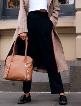 Mulher carregando uma bolsa marrom com calça preta, blusa branca e casaco marrom